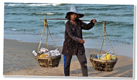 Picture of a woman walking on a beach carrying a stick across her shoulders with two baskets containing goods in opposition to global overconsumption. 