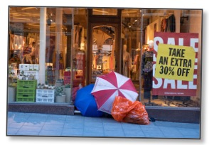 Picture of shop front displaying sale signs with two large open umbrellas and a bag of belongings in the entry as sign of contradiction and global overconsumption. 