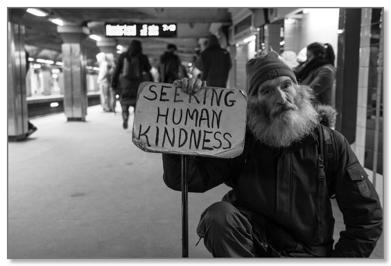 Black and white picture of a man in a mall holding a sign communicating ‘seeking human kindness’ in contrast to mass intolerance and global overconsumption. 