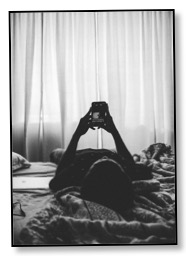 Black and white picture of a person lying alone on a bed looking at a mobile phone in a time of global overconsumption. 