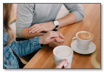 Picture of a couple at a table with hot drinks; one resting a comforting hand on top of the other, communicating the need for support in relationships when feeling alone.