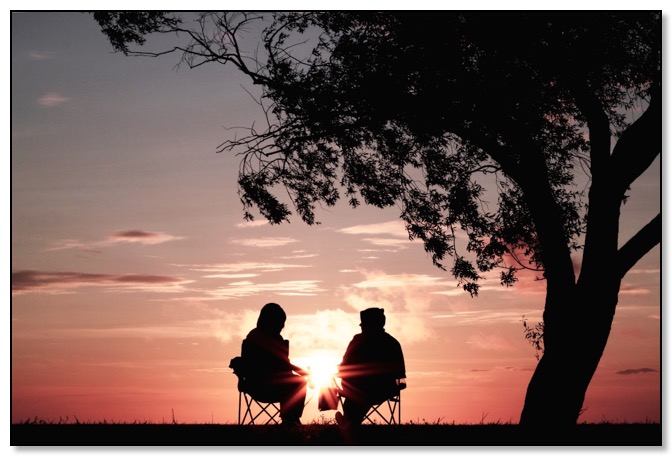 Picture of two people sitting on fold up chairs under a tree at sunset, supporting each other and having relationships.